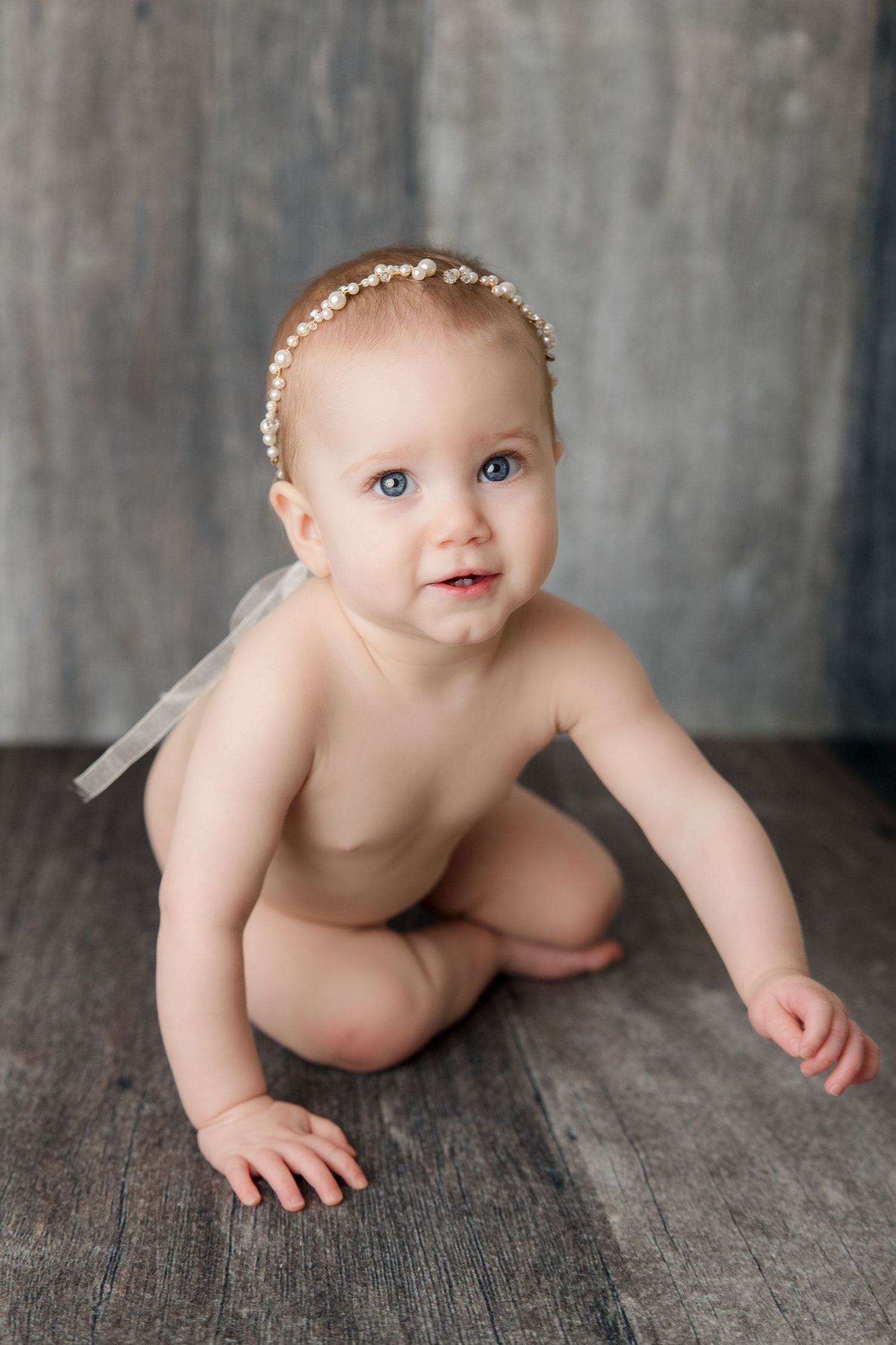 Sweet baby crawls across rustic wooden floor during milestone portrait session wearing delicate white hair bow.