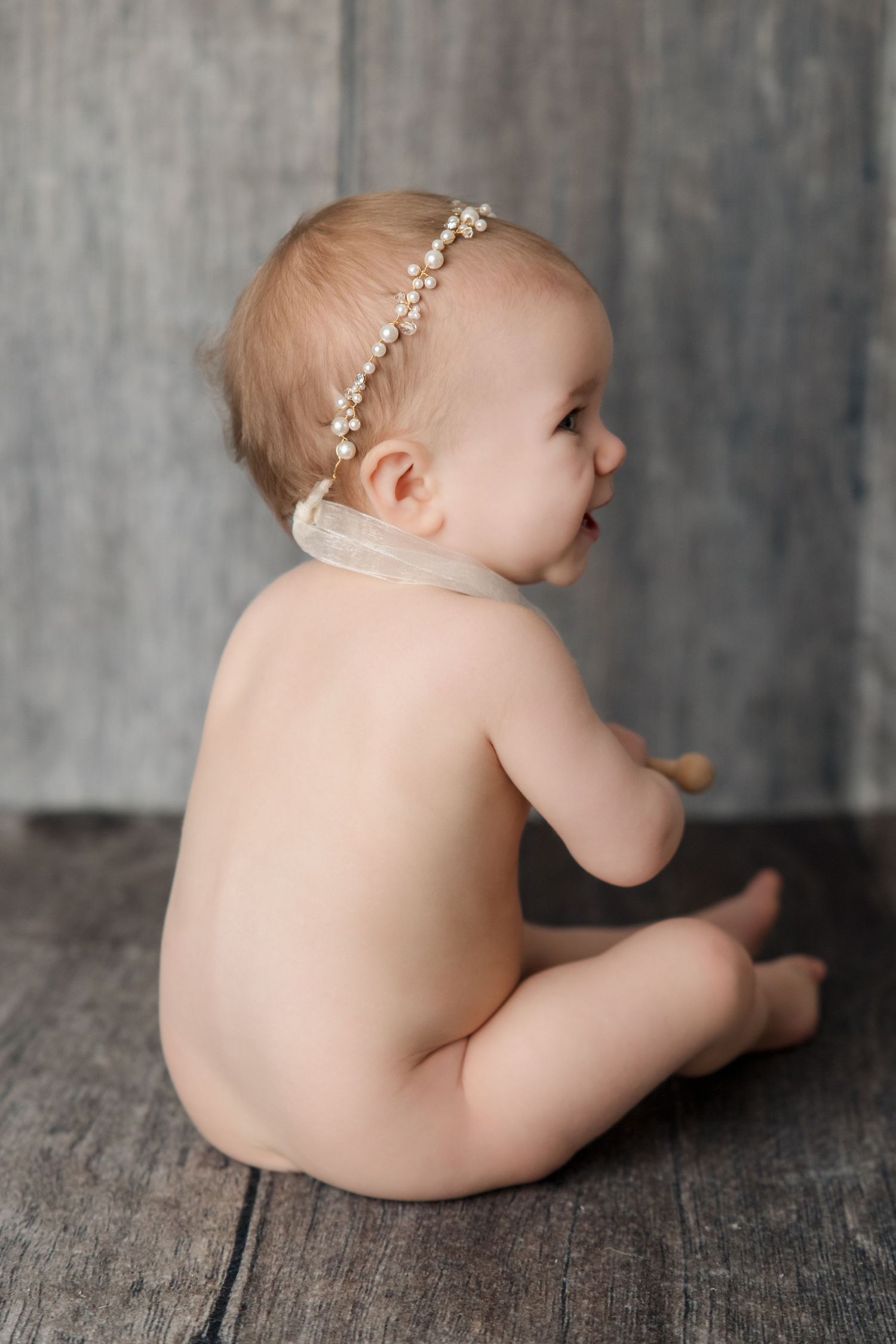 Side profile of baby wearing pearl headband sitting on rustic wood floor against gray backdrop.