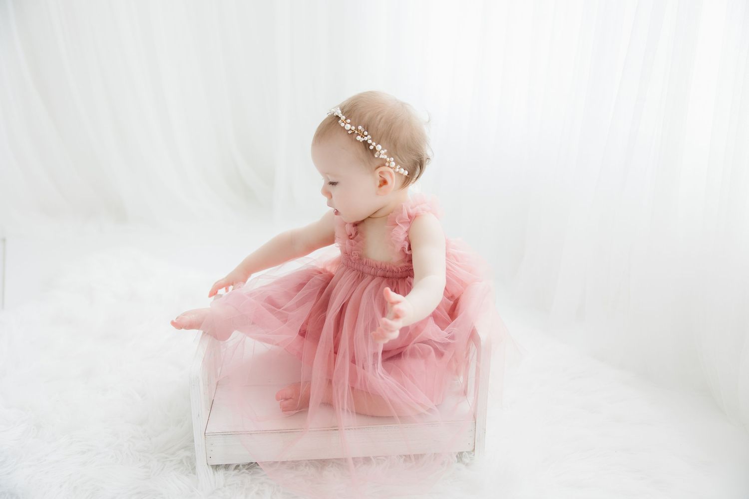 A baby in a pink tutu dress poses elegantly against a soft white background in a ballet-inspired photoshoot.