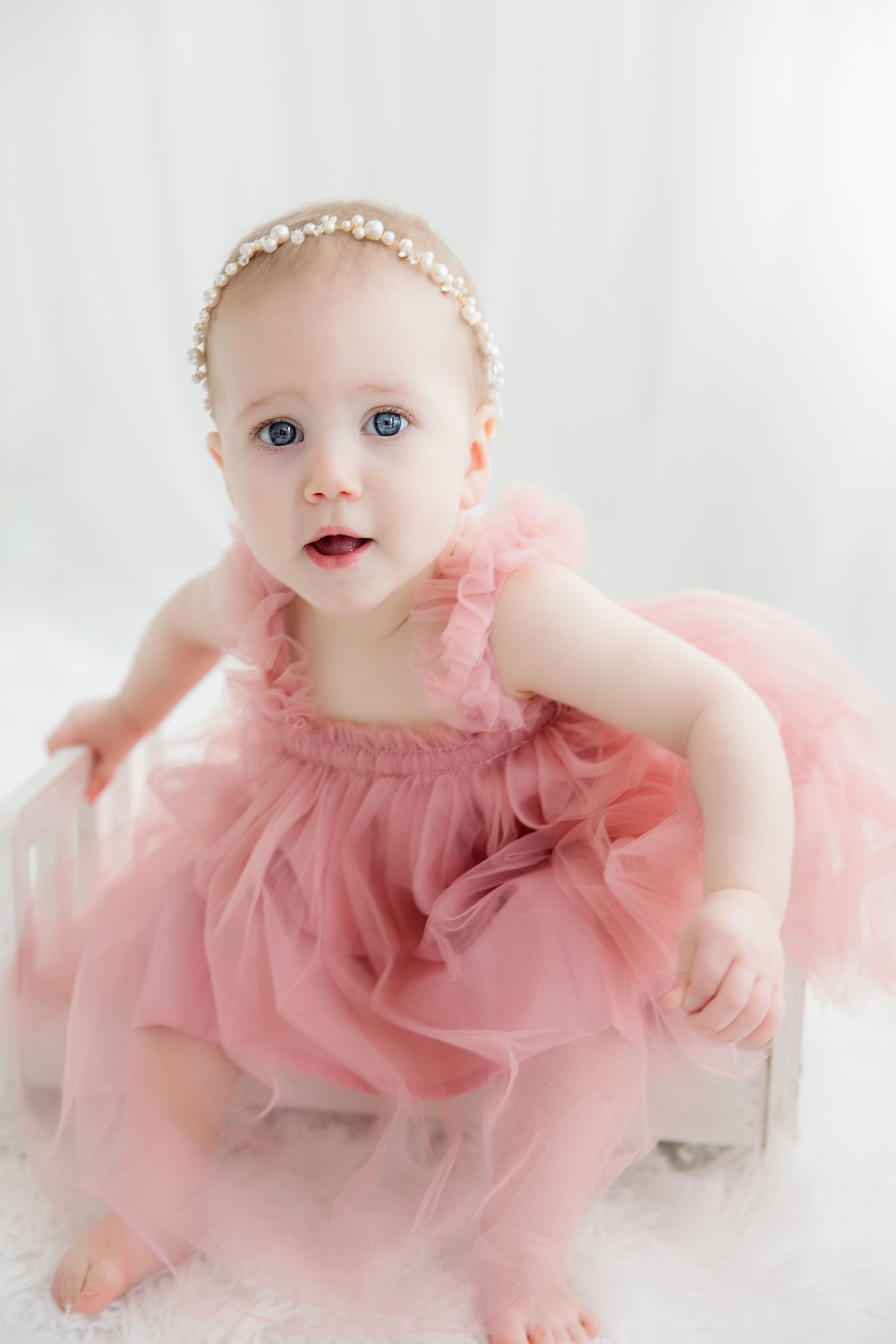 Baby wearing a delicate pink tulle dress and pearl headband poses on white backdrop.