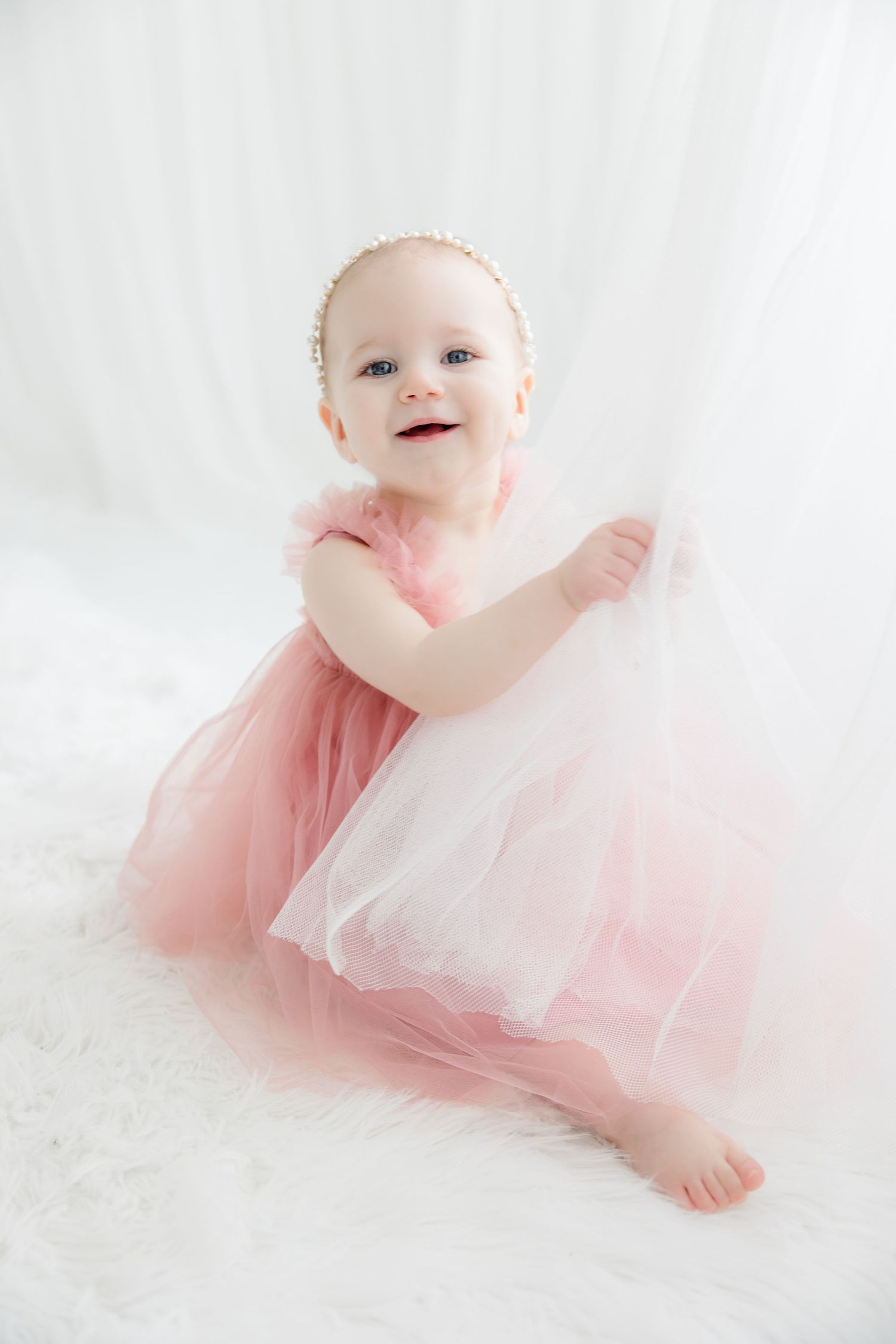 Baby in flowing pink tulle dress and pearl headpiece smiles against white background.