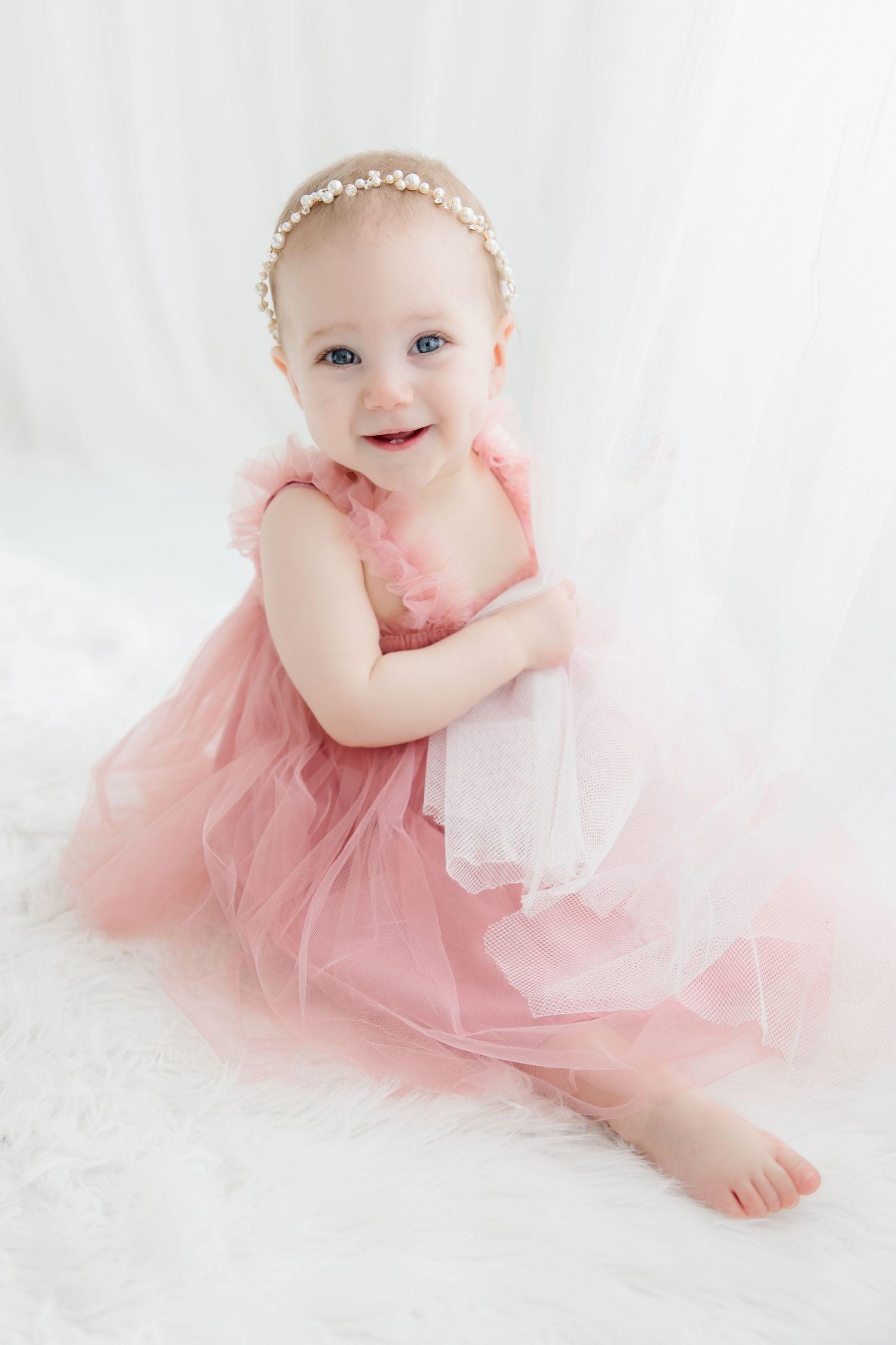 Sweet baby in pink tulle dress and floral headpiece lays on white fluffy backdrop during professional photoshoot.
