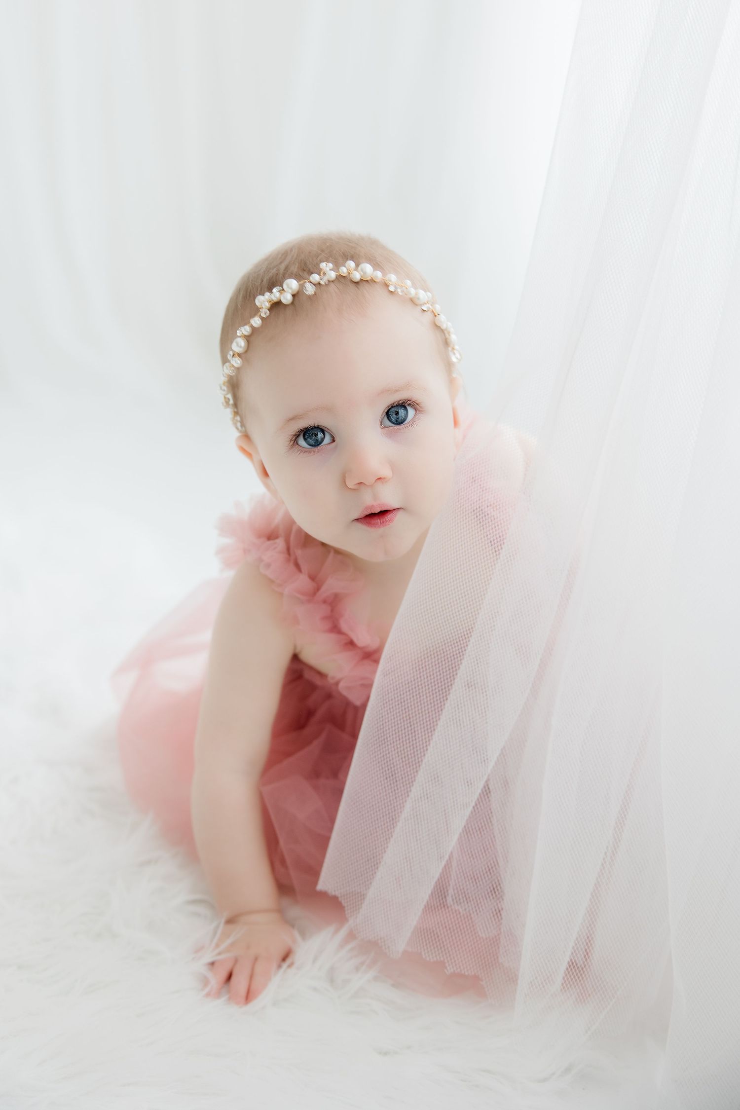 A sweet baby in a pink tulle dress and delicate headband crawls on a soft white backdrop during a photo session.
