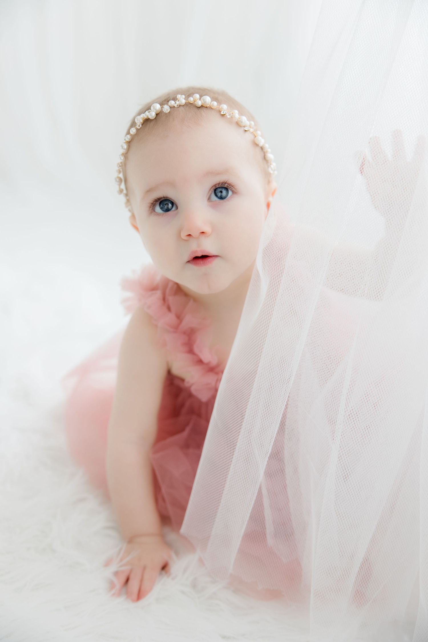 Sweet baby in pink tulle dress and pearl headband lies on soft white fabric backdrop.