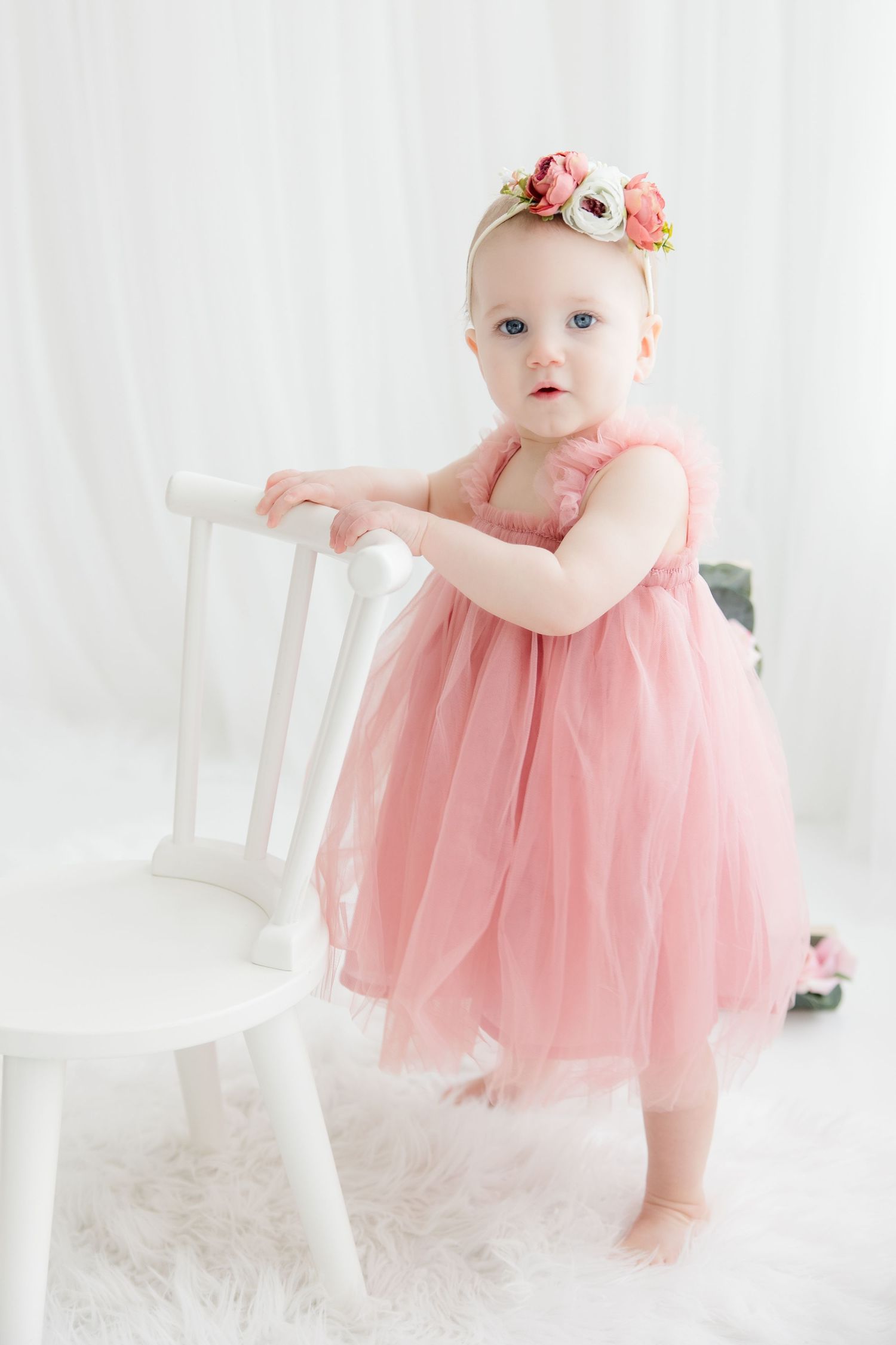 Baby in pink tulle dress holds onto white chair while standing on plush carpet in bright studio setting.