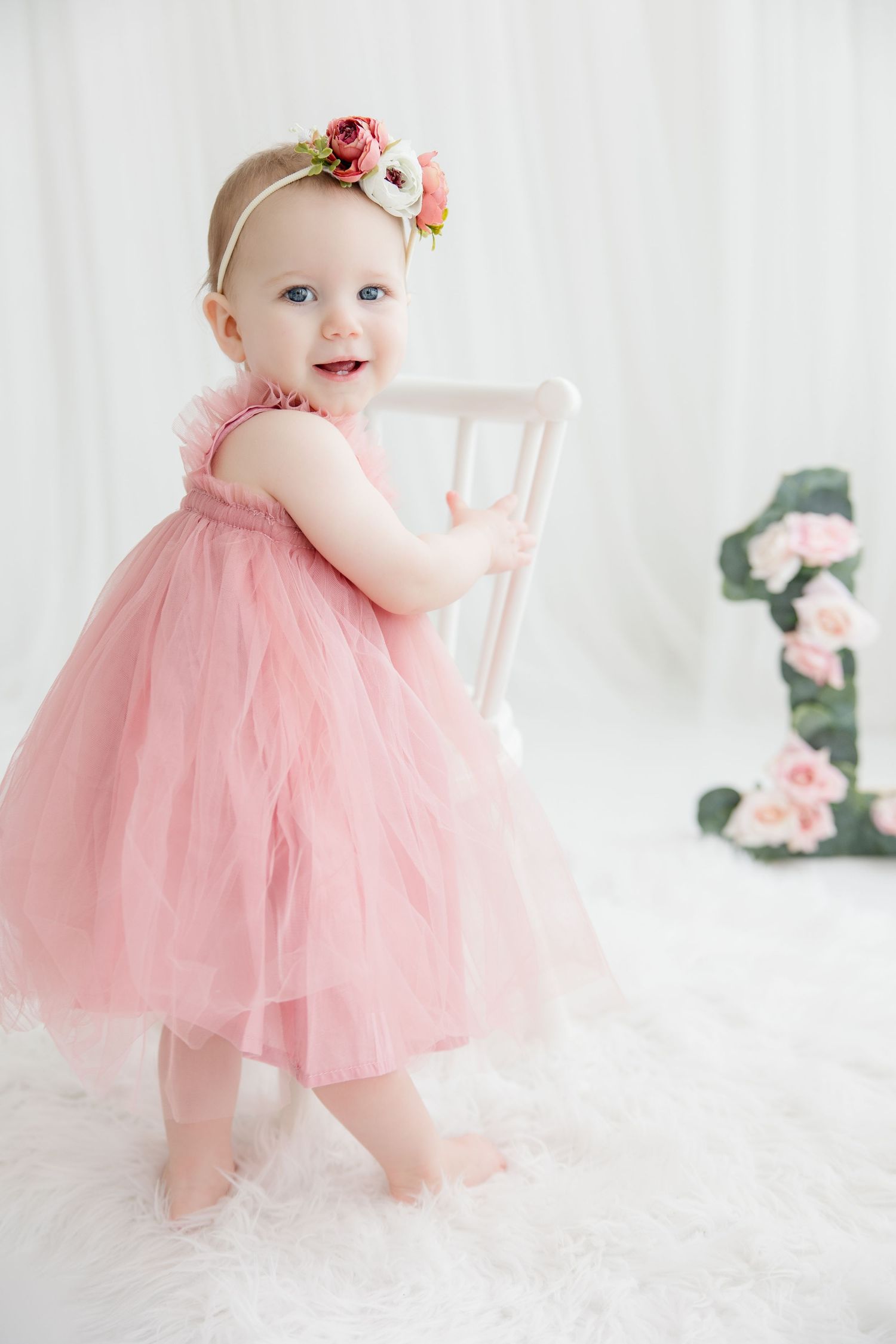 Baby in pink tulle dress poses for professional birthday photoshoot with floral decorations.