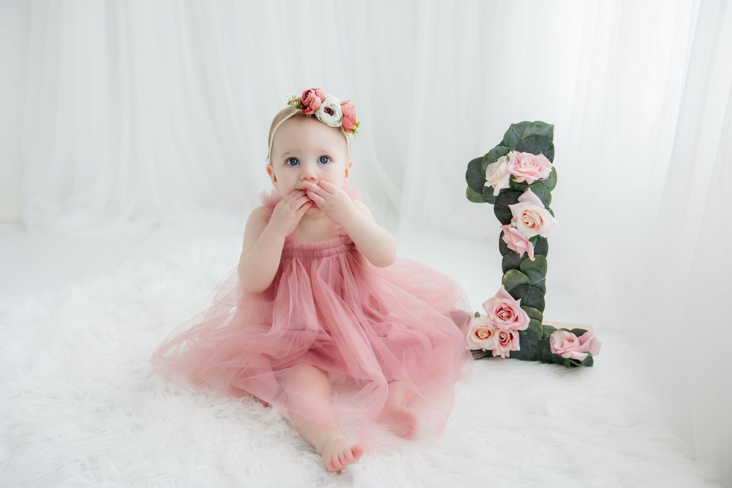 Baby in pink tulle dress crawls on white backdrop during first birthday cake smash photo session.