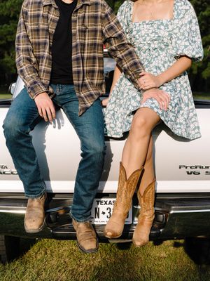 A couple wearing boots sits casually on the edge of a white pickup truck bed against a green outdoor backdrop.