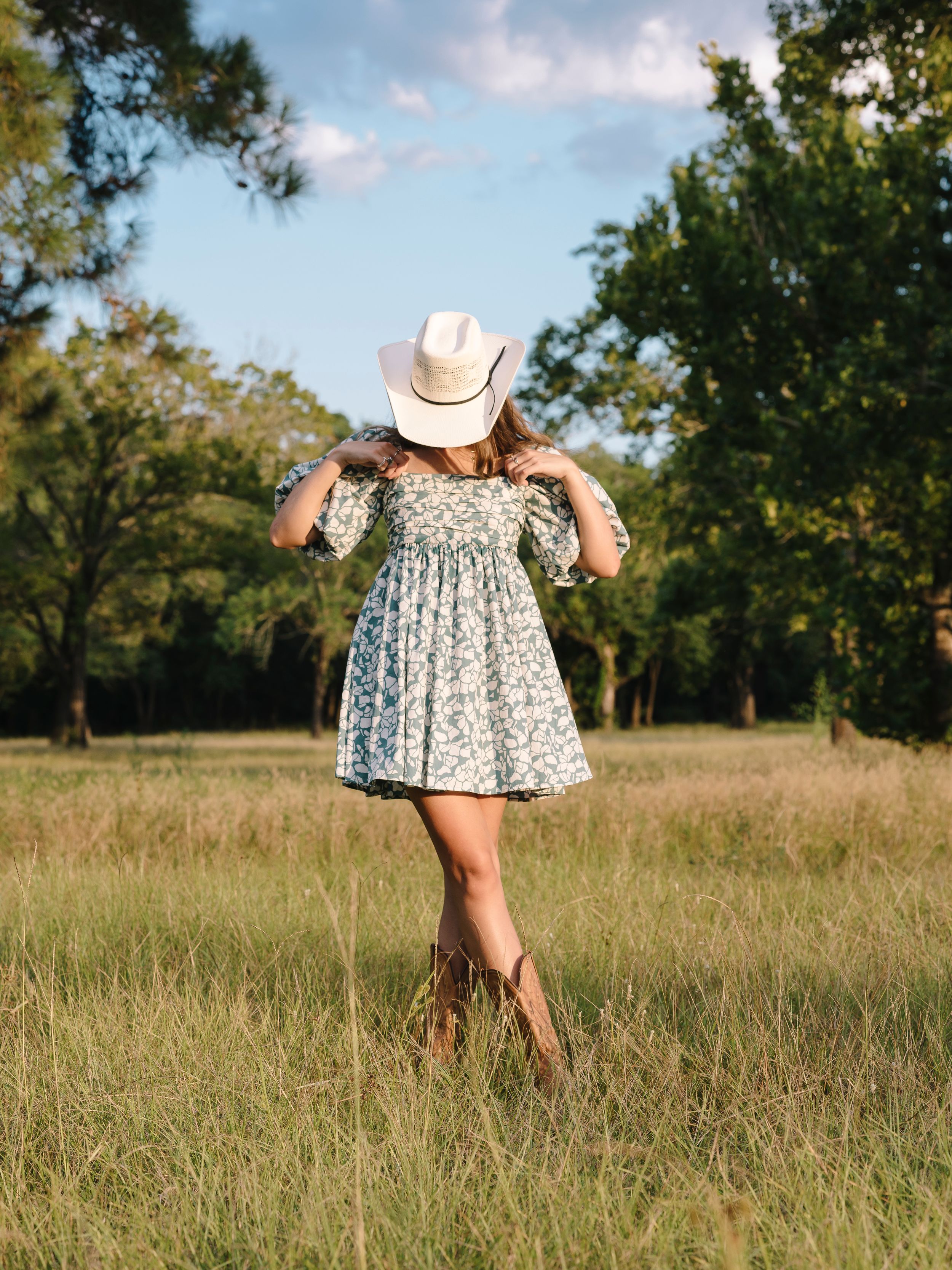 A person in a floral skirt and cowboy hat walks through a sun-dappled field on a warm summer afternoon.