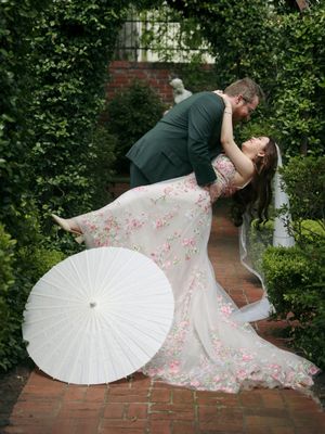 Romantic dance pose in a garden setting with floral dress and white parasol making elegant shadows in natural light.