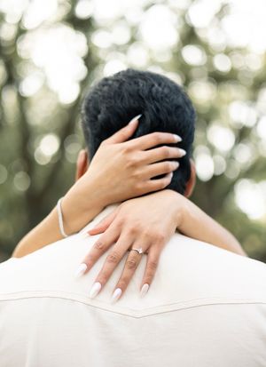 A hand gently rests on a shoulder clothed in a white garment, with soft bokeh lights in the background.