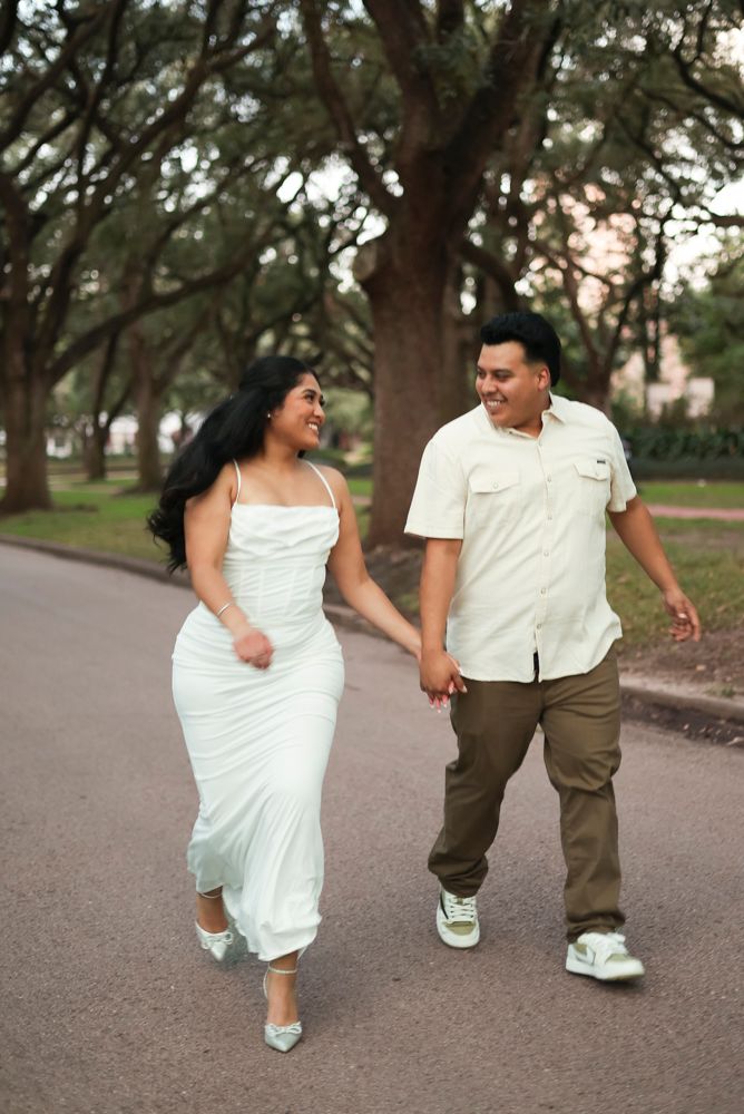 A couple playfully walks and kicks together on a tree-lined path wearing white and brown outfits.