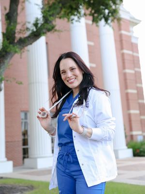 Healthcare professional in blue scrubs and white coat standing in front of brick building with white columns.