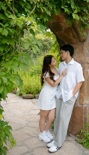 A couple in white outfits shares a romantic moment in a garden walkway with stone walls and green foliage.