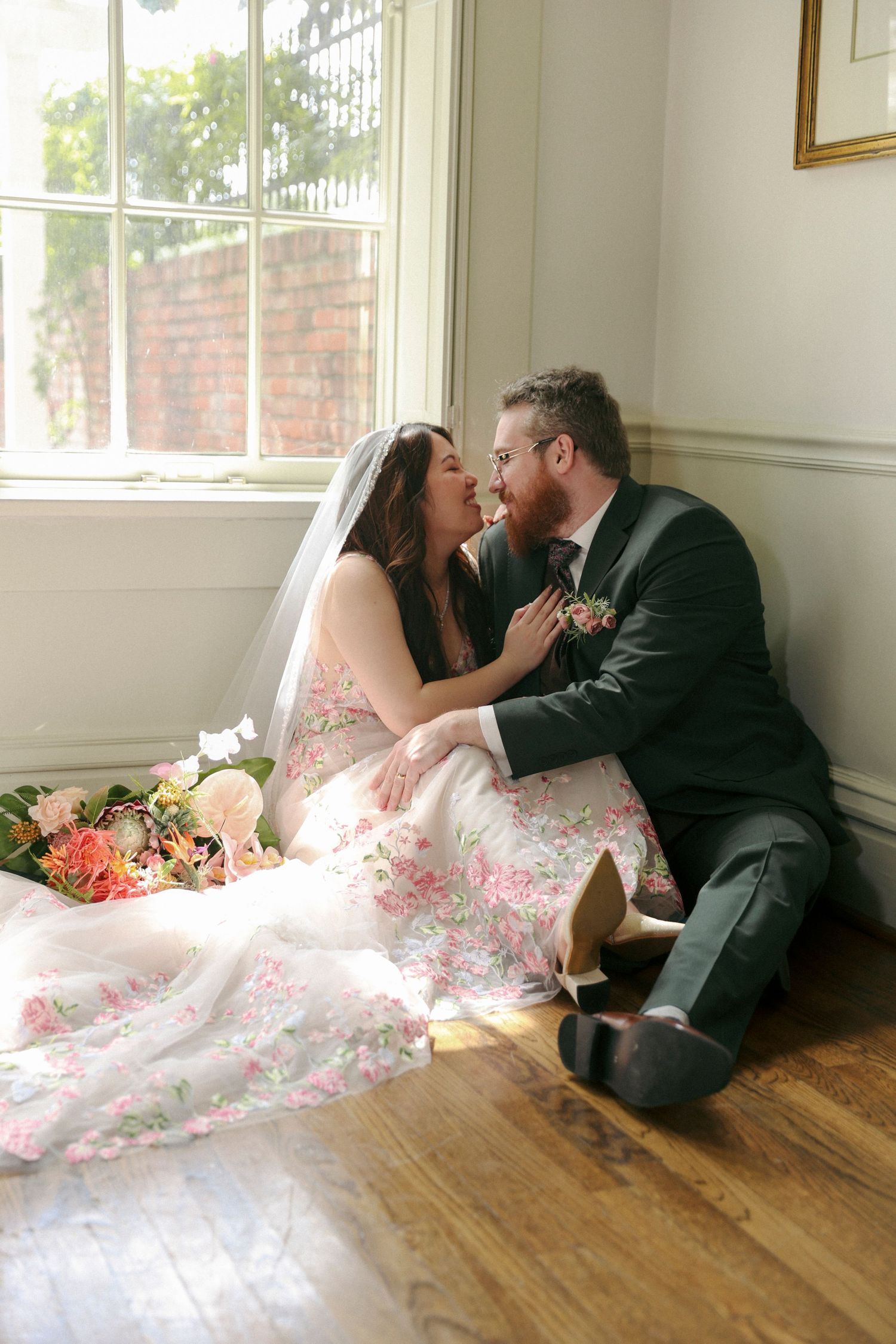 A couple in wedding attire share an intimate moment while sitting on hardwood floors near a sunny window.
