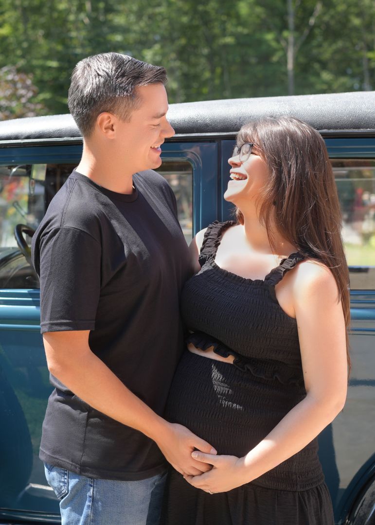 A couple in black clothing embraces and smiles while standing next to a vintage blue car outdoors.