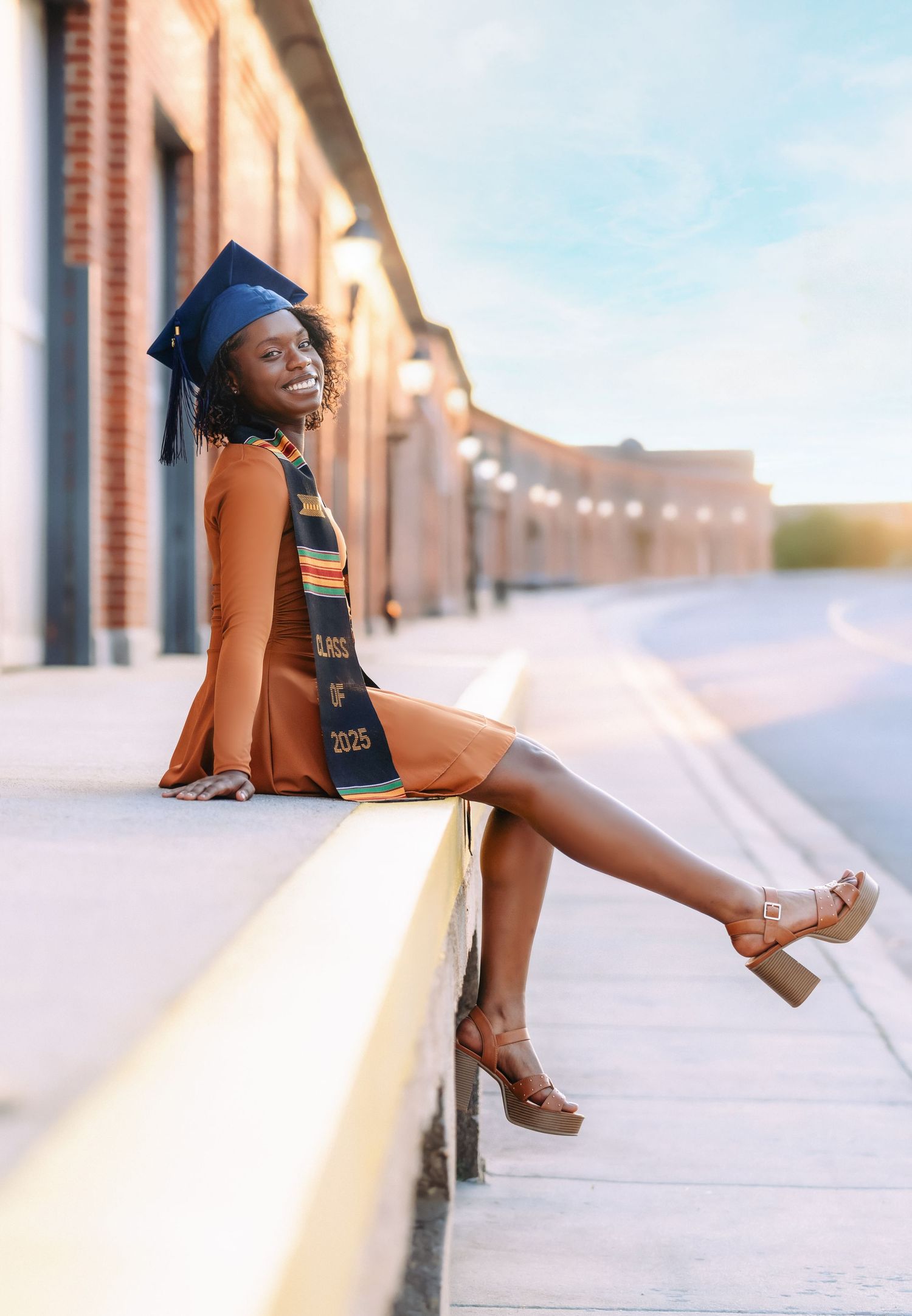 Smiling graduate in cap and Kente stole posing on ledge in golden hour senior portrait.