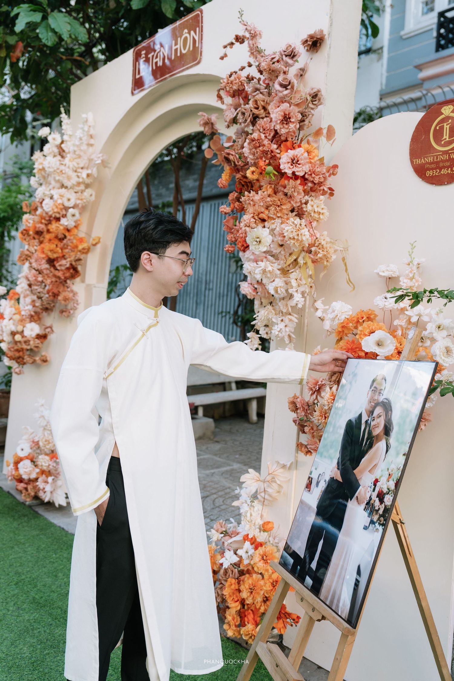 Groom in white ao dai touches wedding photo on easel by floral arch with orange blooms.