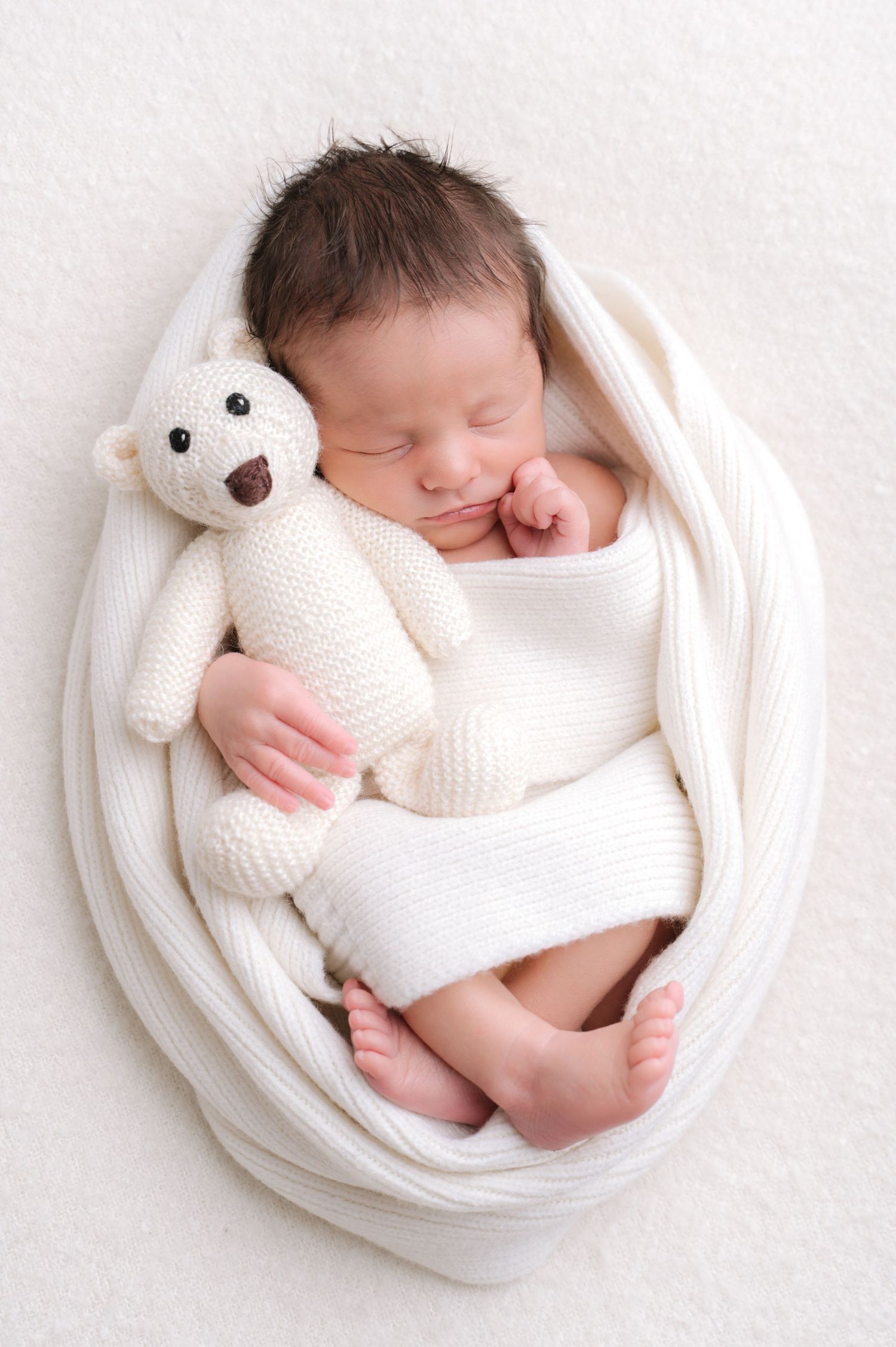 Baby in white outfit sleeping next to knitted teddy bear.