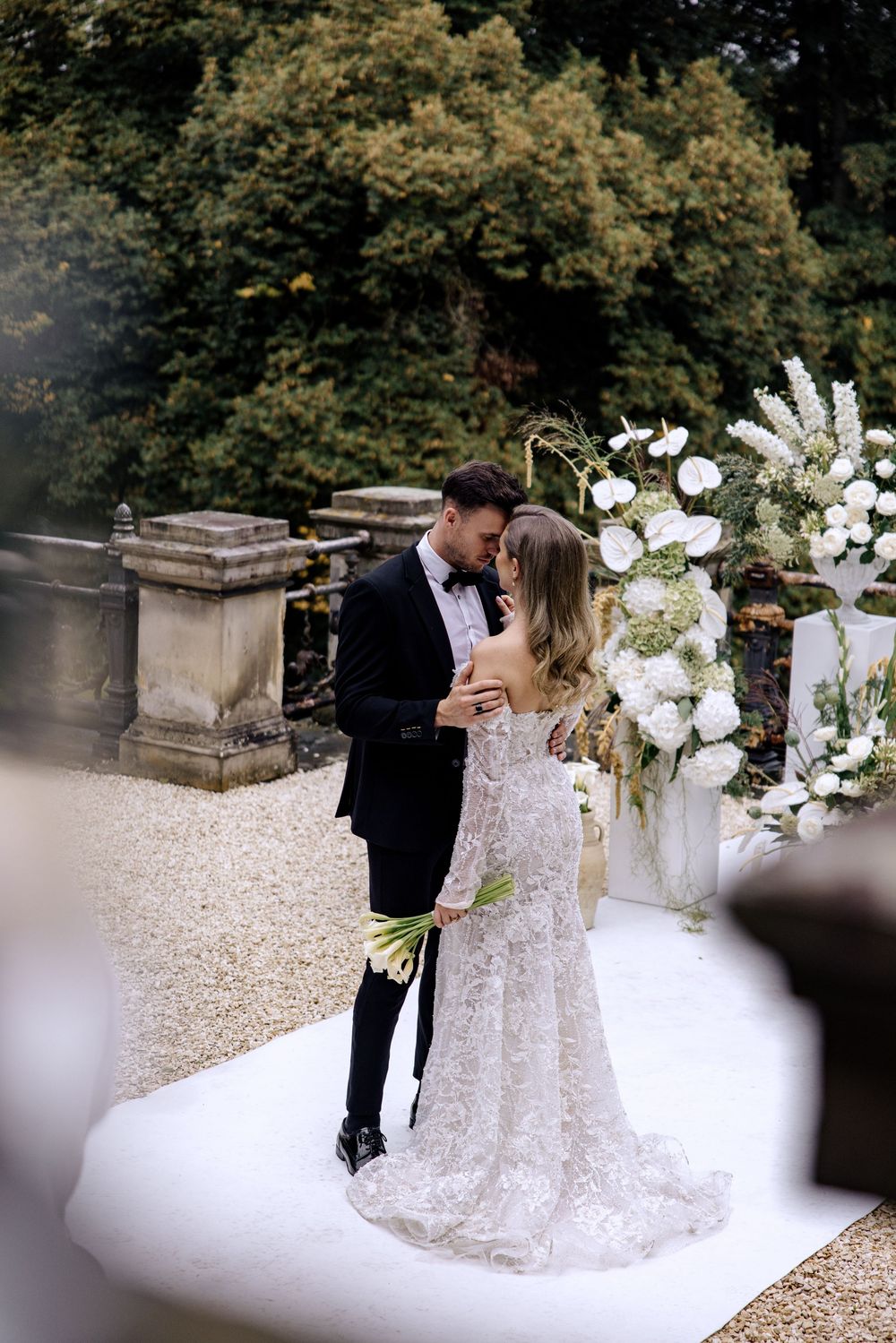 A romantic wedding couple embraces on a gravel path surrounded by lush greenery and elegant white floral arrangements.