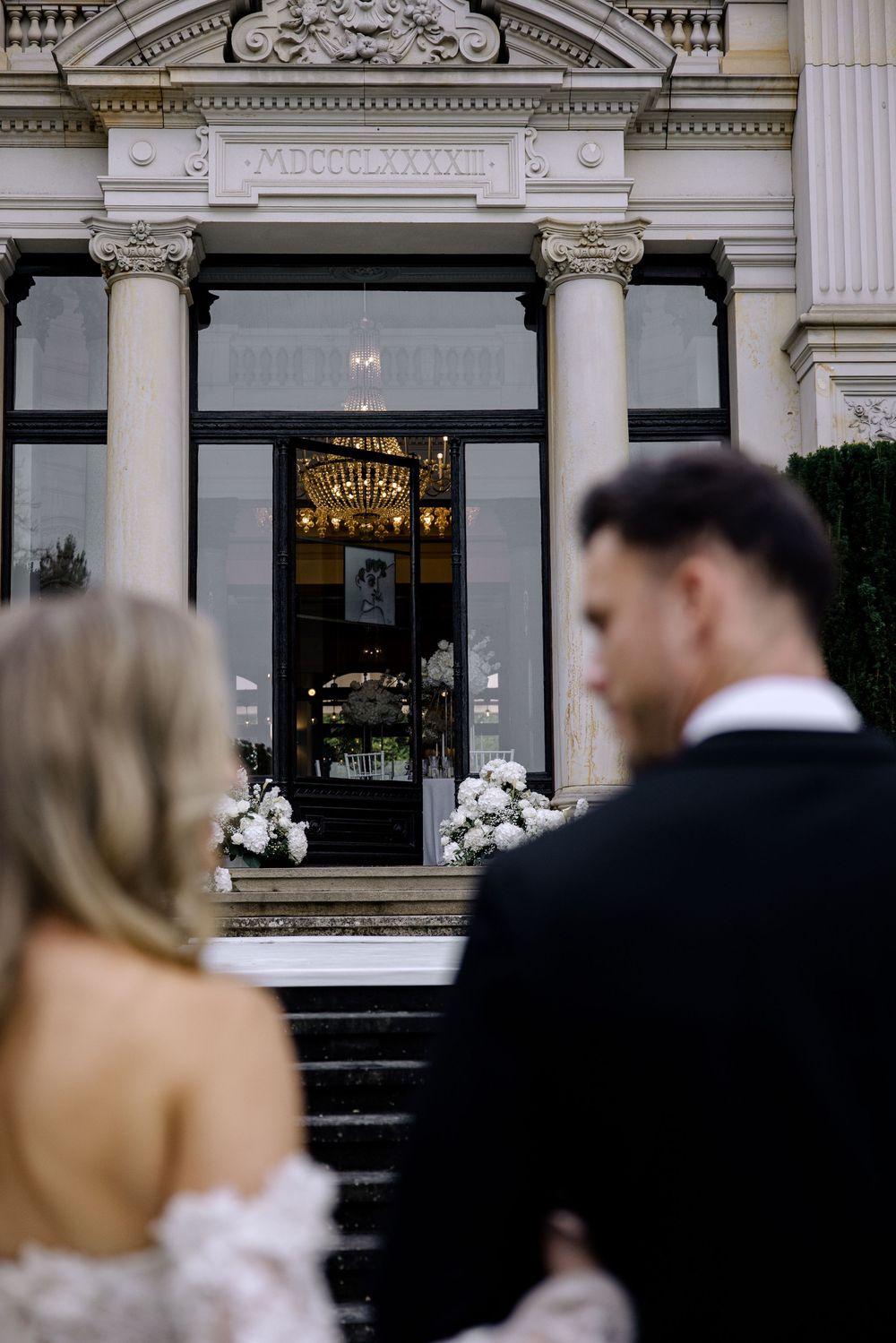 A wedding couple walks past an ornate historic building with elegant architectural details and large windows.