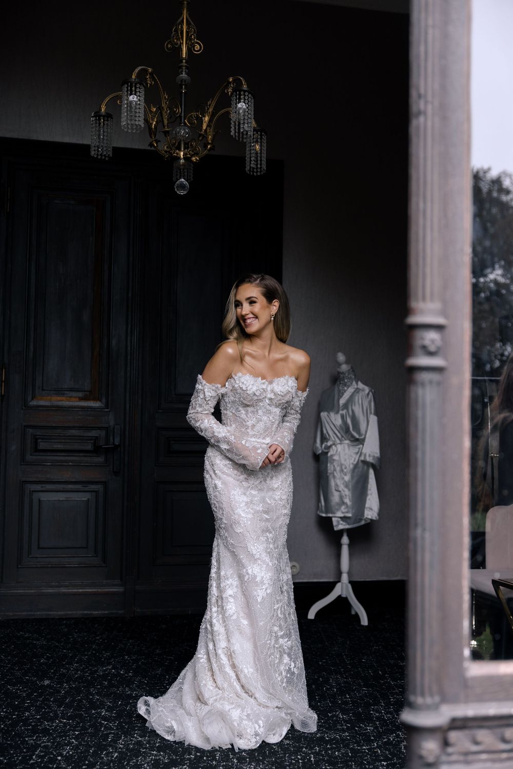 Bride in off-shoulder lace wedding gown poses elegantly in front of dark doorway with chandelier overhead.