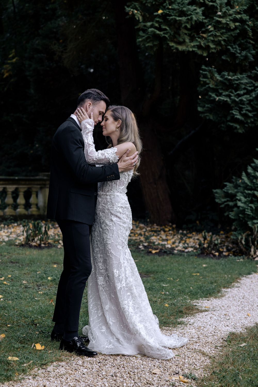 A romantic wedding couple shares a kiss on a gravel path surrounded by lush trees and fallen autumn leaves.