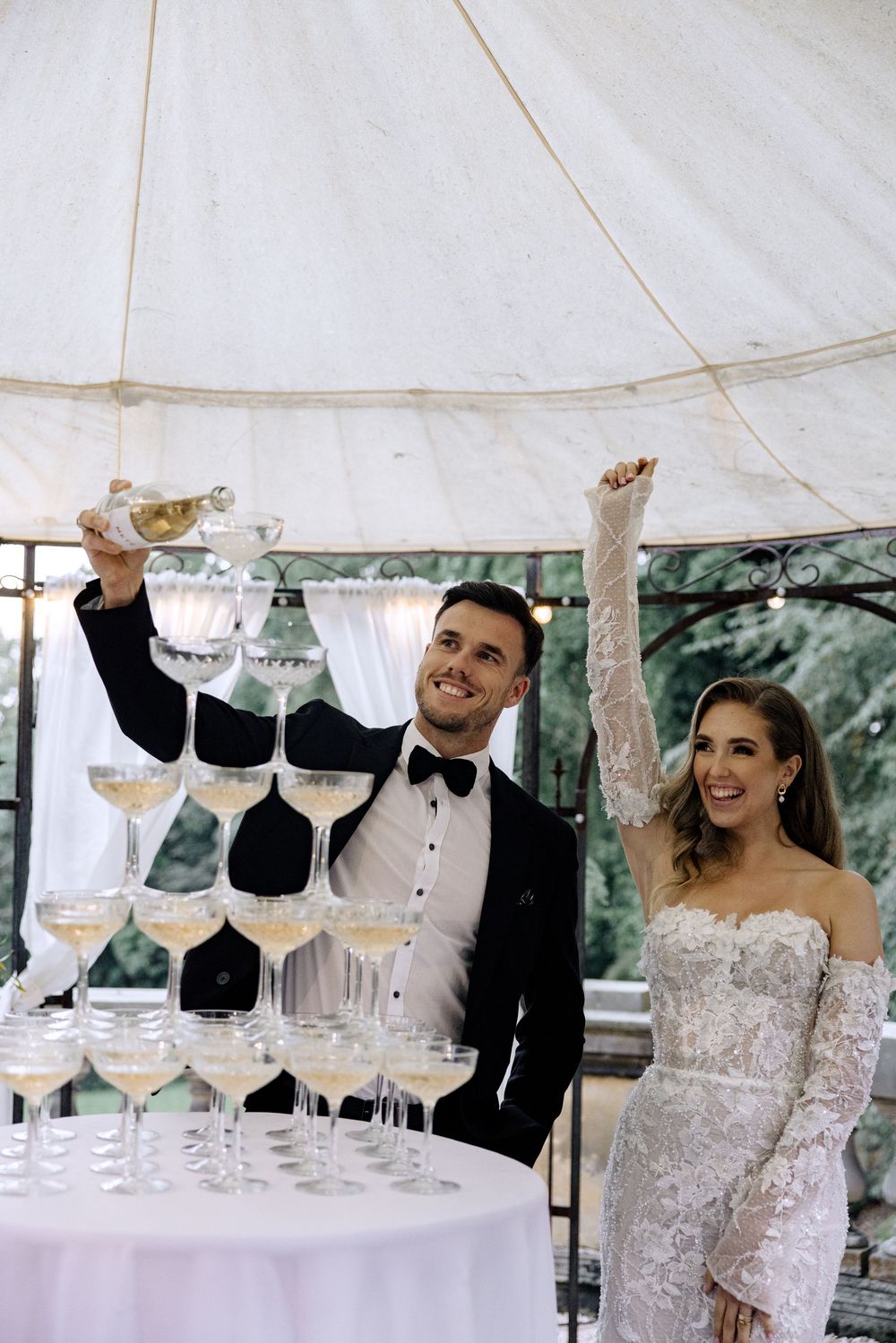 Newlyweds celebrate with a champagne tower under an elegant white tent at their wedding reception.
