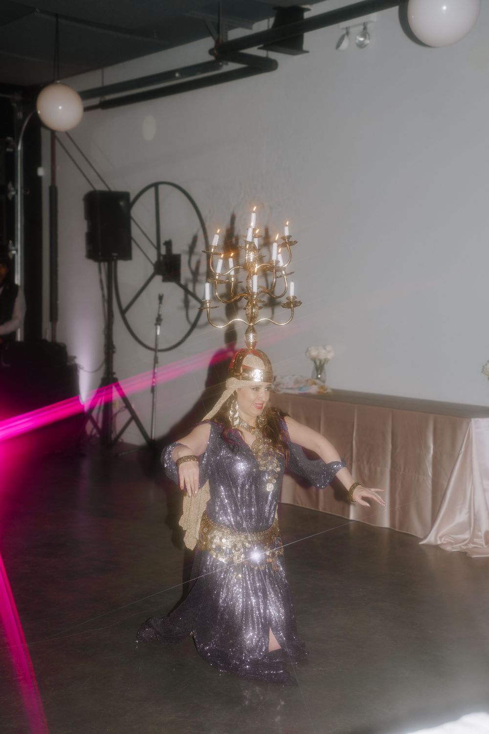 A person in a sparkly silver dress dances on an empty dance floor with elegant chandeliers hanging above.