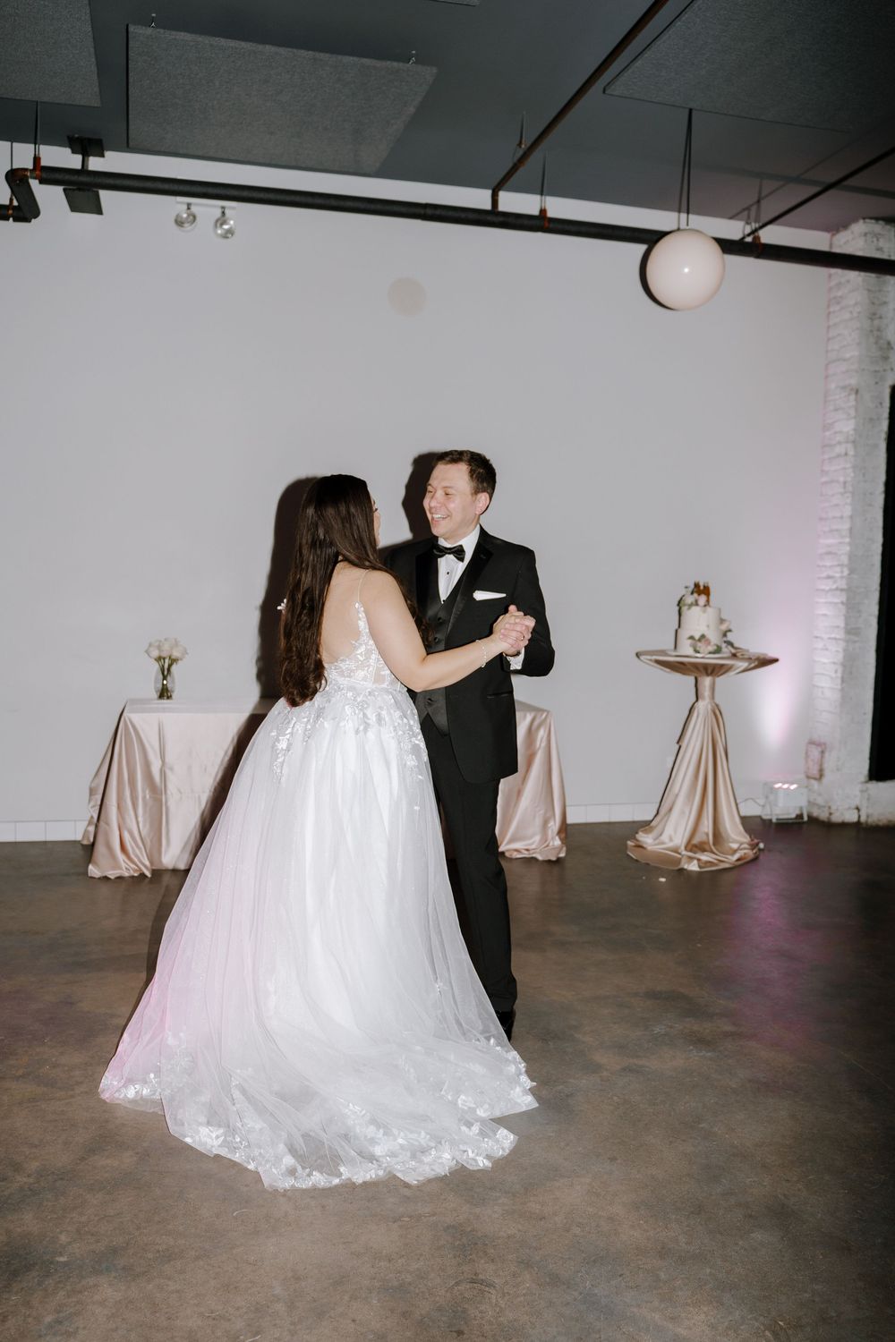 A couple dances together in an industrial venue space with round tables decorated for a wedding reception.