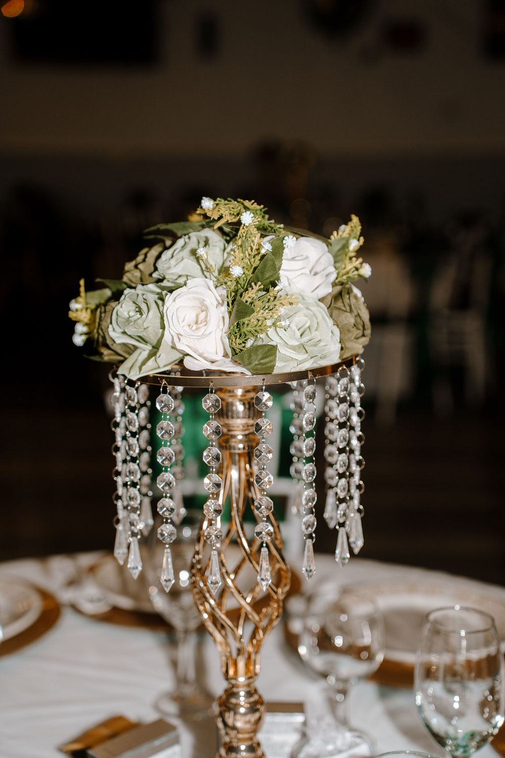 Elegant wedding centerpiece with white roses and crystal drops on a gold candlestick holder atop a white tablecloth.