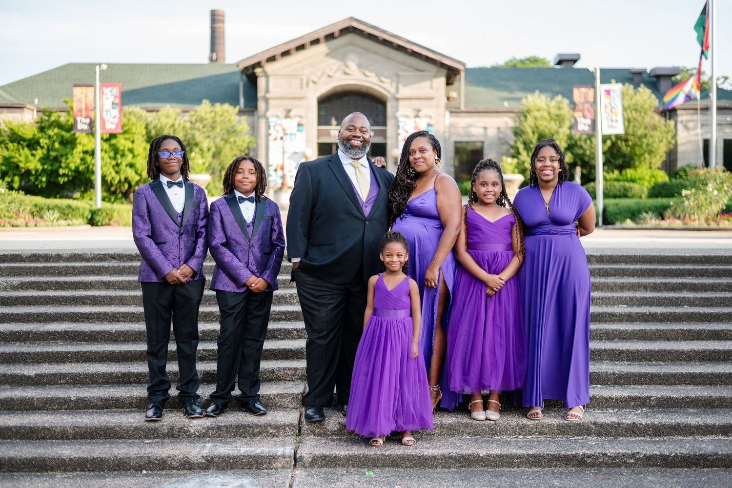Wedding party members in coordinated purple attire pose together on stone steps in front of a building.