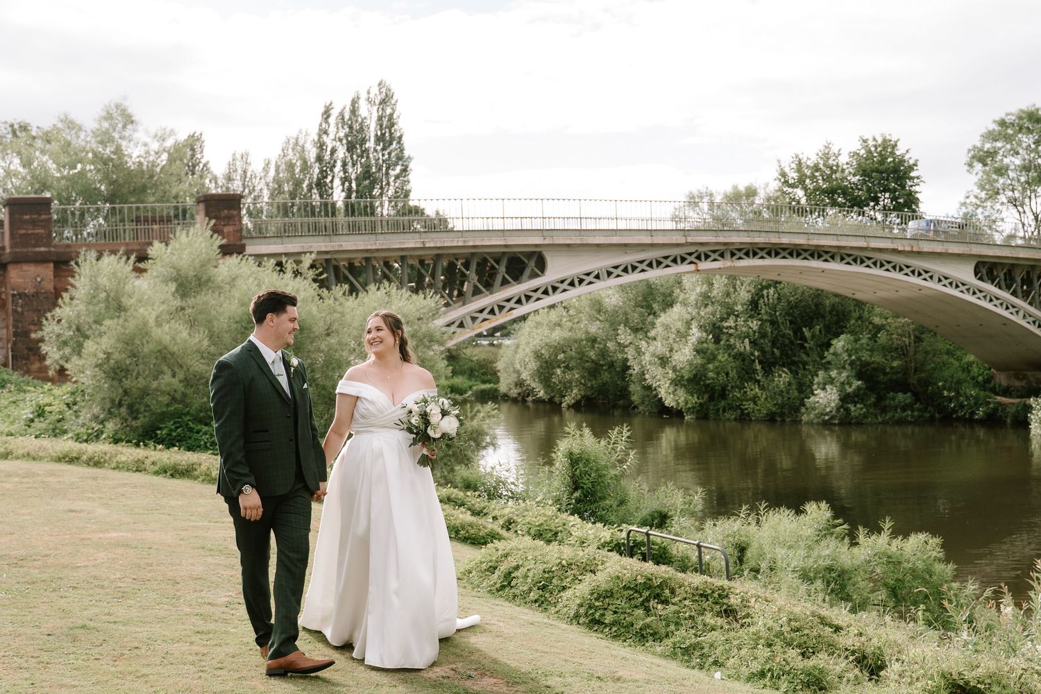 Romantic couple strolls along a scenic riverbank with an ornate bridge in the background during a wedding photoshoot.