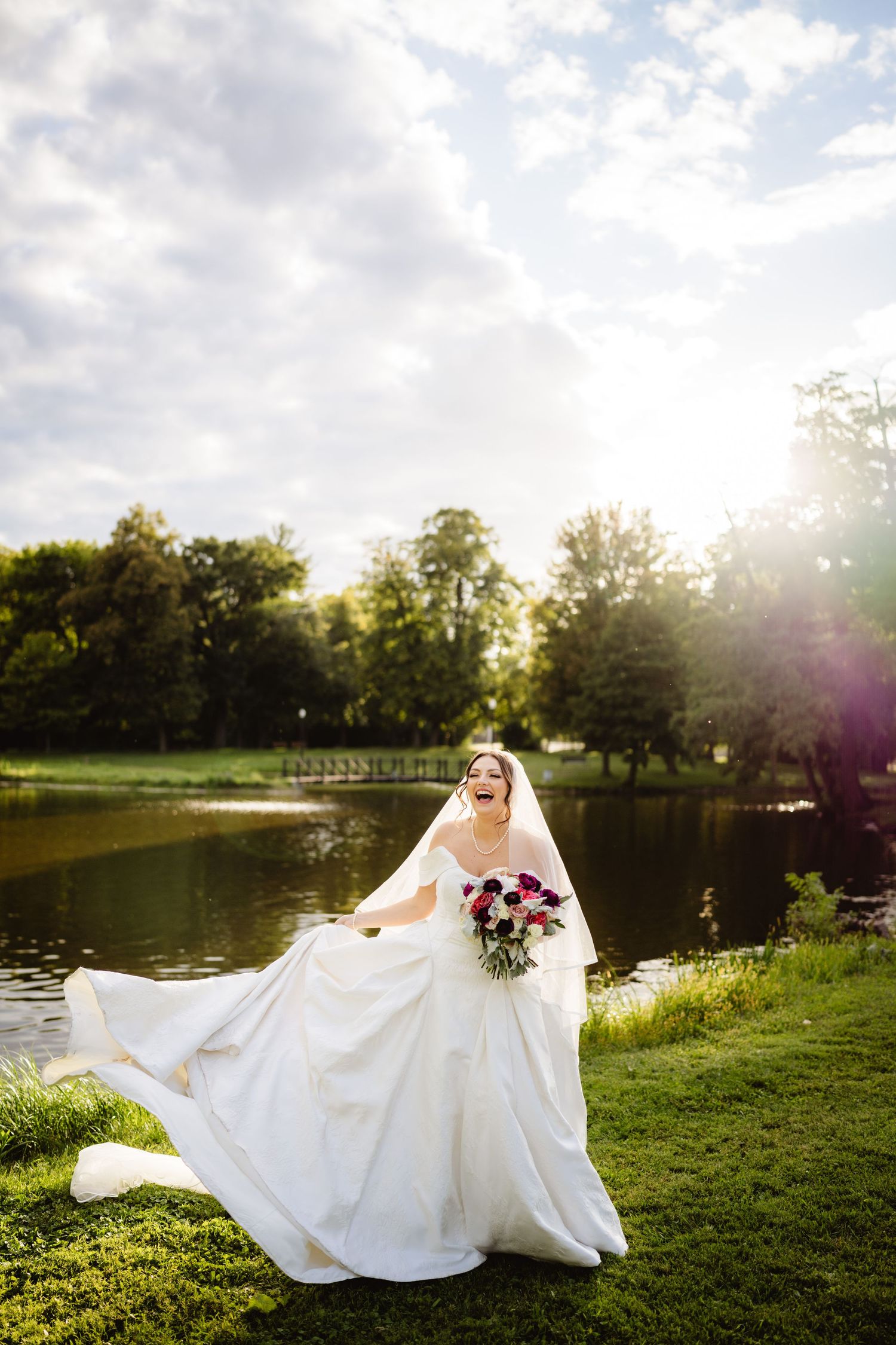 Bride in flowing white wedding dress poses gracefully by a serene lake at sunset creating magical bridal portraits.