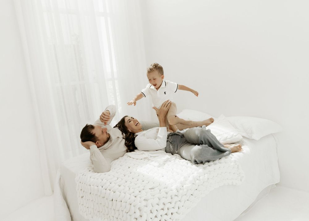 Parent and baby playing together on a white bed with textured bedding in a minimalist bright setting.