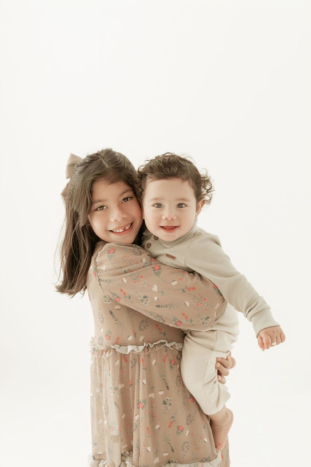 Two siblings in neutral-toned clothing embrace and smile against a white background in a heartwarming portrait.