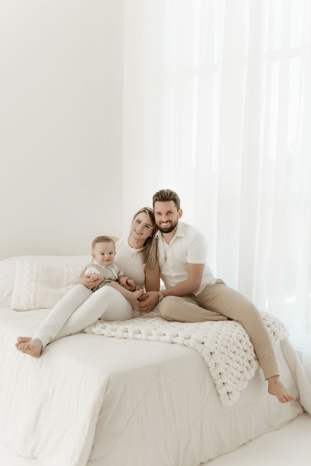 A family relaxes together on a white bed wearing neutral colors in a bright, airy bedroom setting.