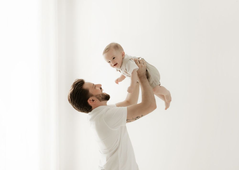 A parent playfully lifts a baby wearing white clothing against a bright white background in a series of photos.