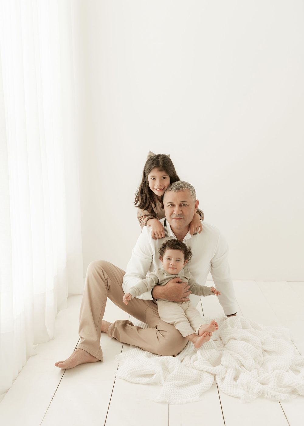 A family wearing white clothing sits together on a white floor in a bright, minimalist setting.