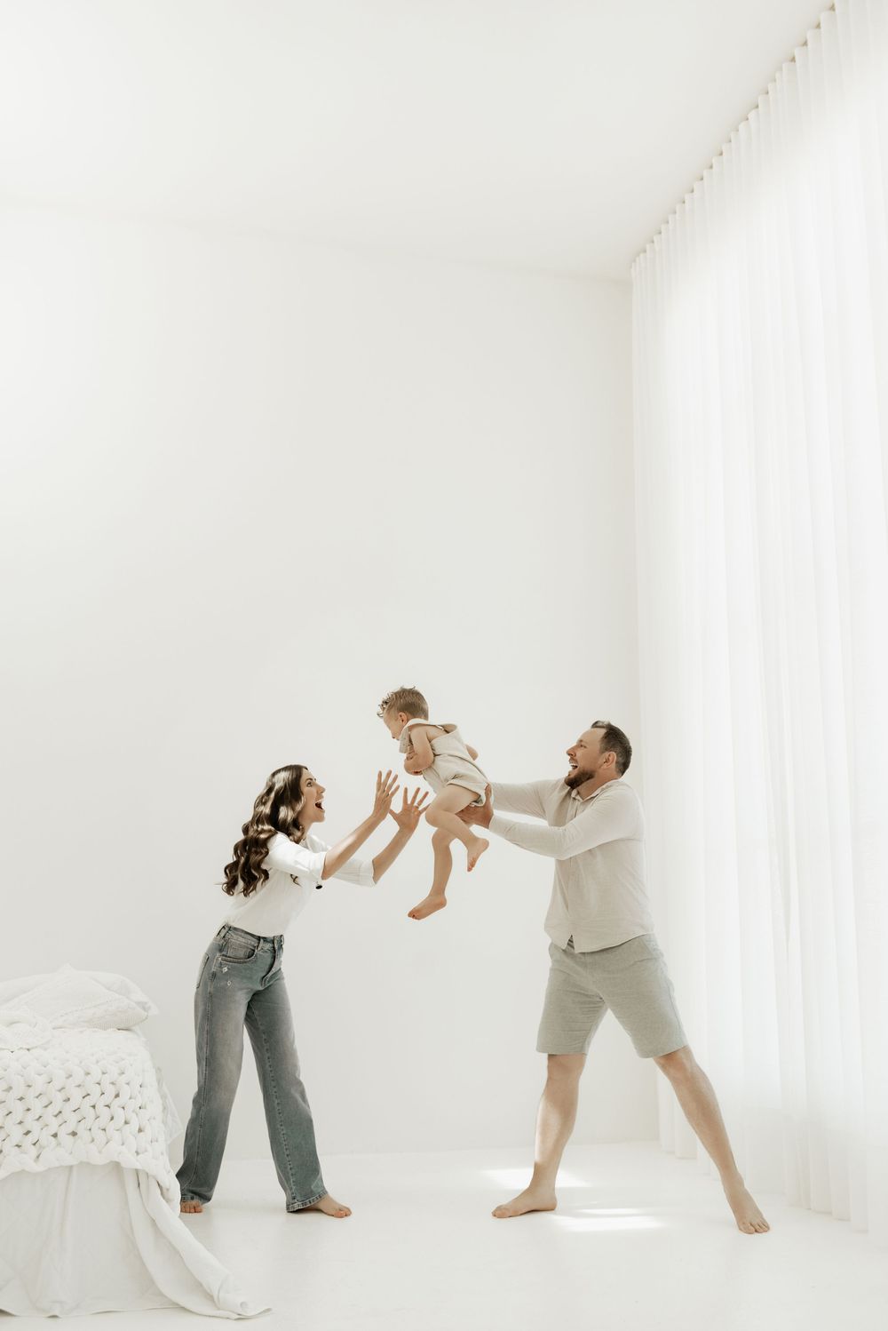 Family plays together in a bright white minimalist bedroom, lifting child playfully in the air.