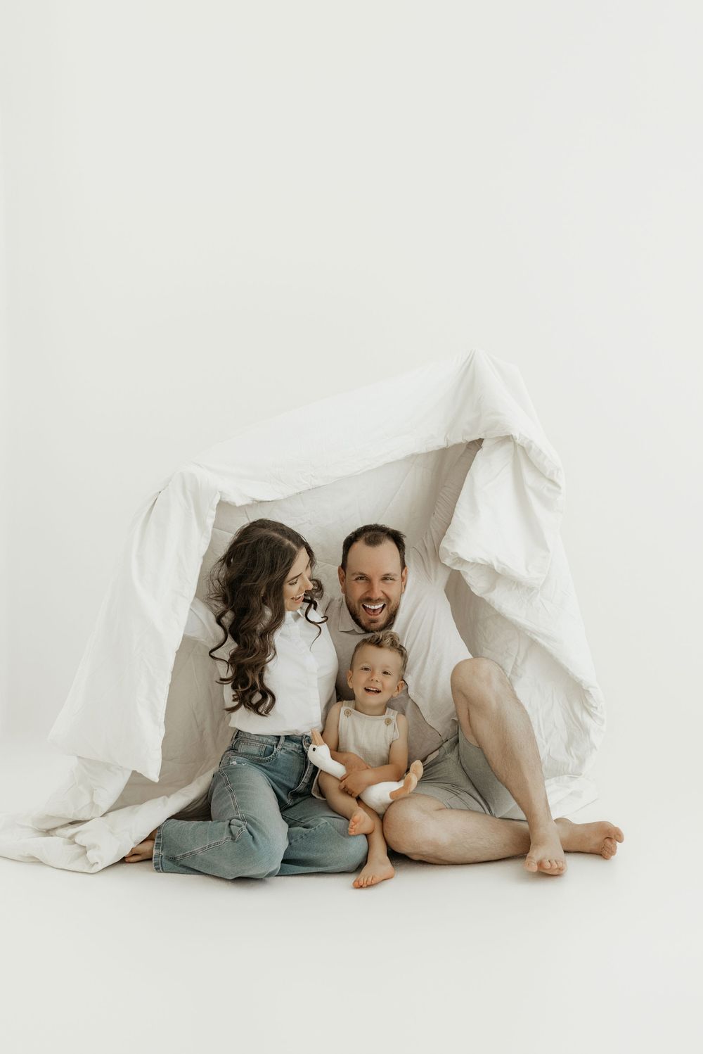 A parent and child share playful moments under white bedsheets in a minimalist photo series.