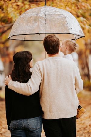 A group huddled together under a clear umbrella walks along an autumn path with golden fall foliage in the background.