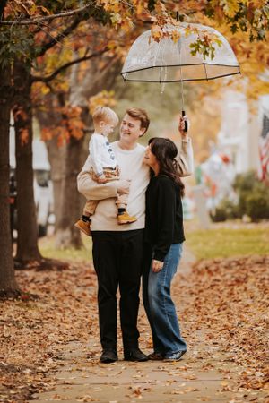 Family shares a tender moment under a clear umbrella surrounded by falling autumn leaves in a park setting.