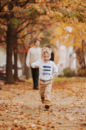 Joyful toddler runs through fallen autumn leaves on a path lined with golden trees while wearing casual clothes.