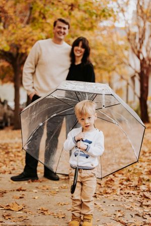 A small child holds a clear umbrella while standing on fallen autumn leaves with people in the background.