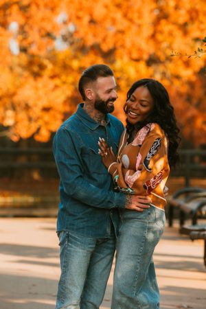 Joyful couple sharing a candid moment of laughter against golden fall trees in casual denim outfits.