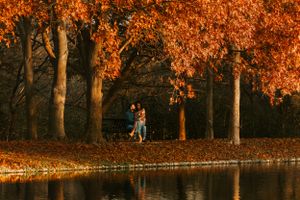 Two people stand under vibrant orange autumn trees beside a tranquil lake reflecting the warm sunset light.