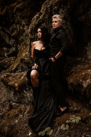 A couple in dramatic black attire poses against a dark rocky cliff in a moody outdoor photoshoot.