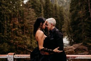 A romantic couple in dark clothing embrace on a bridge surrounded by autumn forest and river scenery.