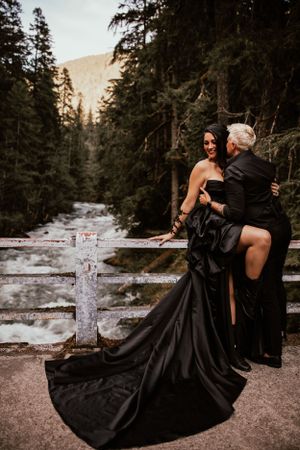 Two people embrace in a romantic scene wearing black attire against a scenic mountain forest backdrop near a river.